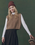A woman with long blonde hair wears The Great Inc.’s Brushed Melange Sweater over a white turtleneck and black skirt, standing by a brown leather chair against a green backdrop.