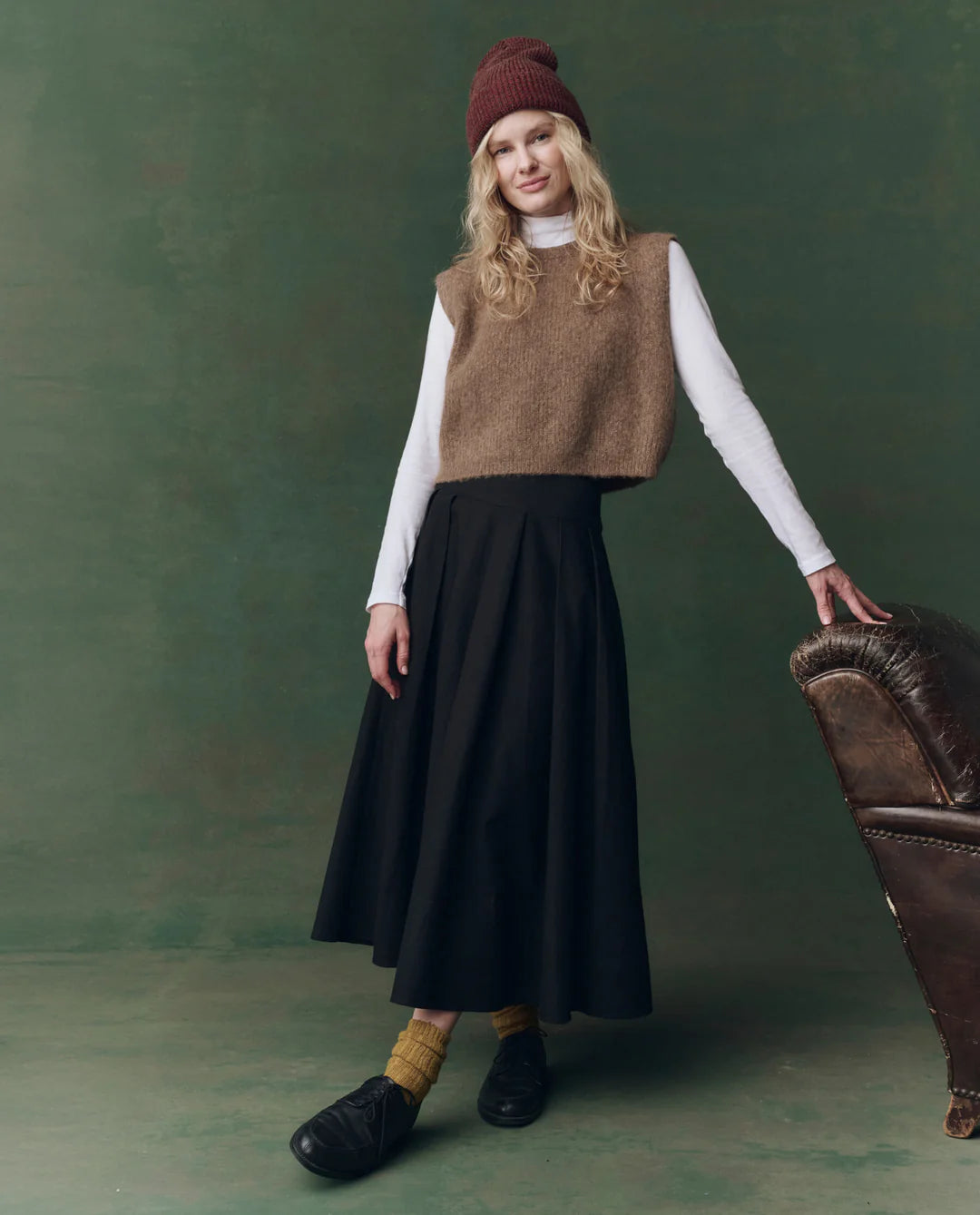 Wearing The Great Inc.'s Brushed Melange Sweater, a person in a white turtleneck, long black skirt, yellow socks, and black shoes leans on a brown leather armchair against a green backdrop.