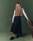 Wearing The Great Inc.'s Brushed Melange Sweater, a person in a white turtleneck, long black skirt, yellow socks, and black shoes leans on a brown leather armchair against a green backdrop.