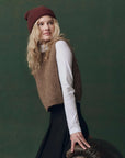 A woman with long blonde hair wears The Brushed Melange Sweater by The Great Inc. over a white long-sleeve shirt and black skirt, posing against a dark green backdrop while leaning on a leather armchair.