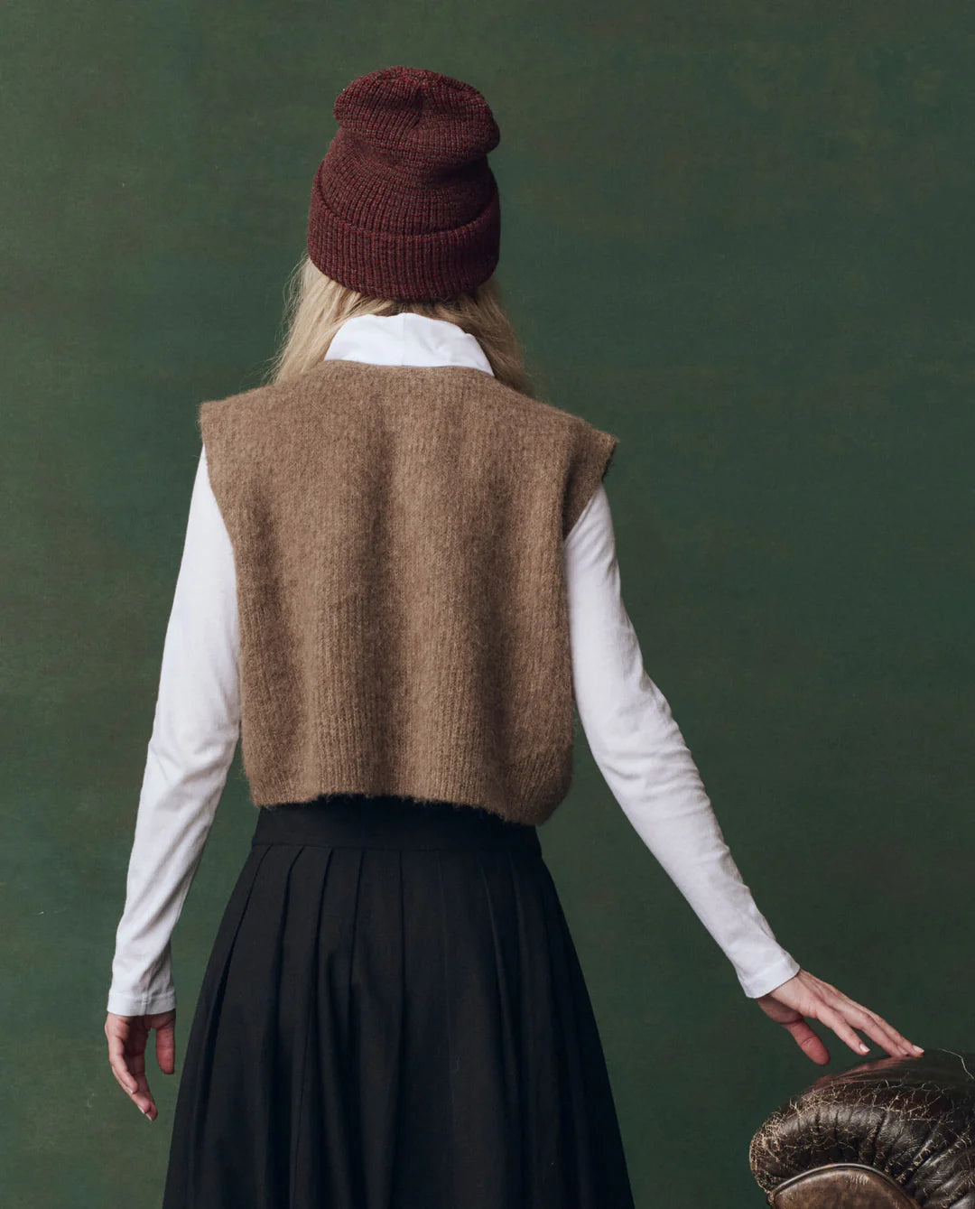 A person wears The Brushed Melange Sweater by The Great Inc. over a white long-sleeve shirt and black skirt, paired with a maroon knit beanie, standing against a dark green background.