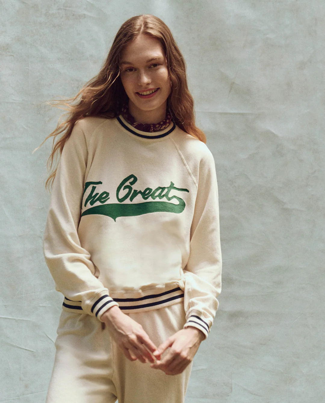 A person wears "The Varsity College Sweatshirt w/ Crest Graphic" by The Great Inc., in oversized cream cotton terry with green text, paired with matching pants. They have long hair and stand smiling in front of a textured, light-colored backdrop.