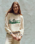 A person wears "The Varsity College Sweatshirt w/ Crest Graphic" by The Great Inc., in oversized cream cotton terry with green text, paired with matching pants. They have long hair and stand smiling in front of a textured, light-colored backdrop.