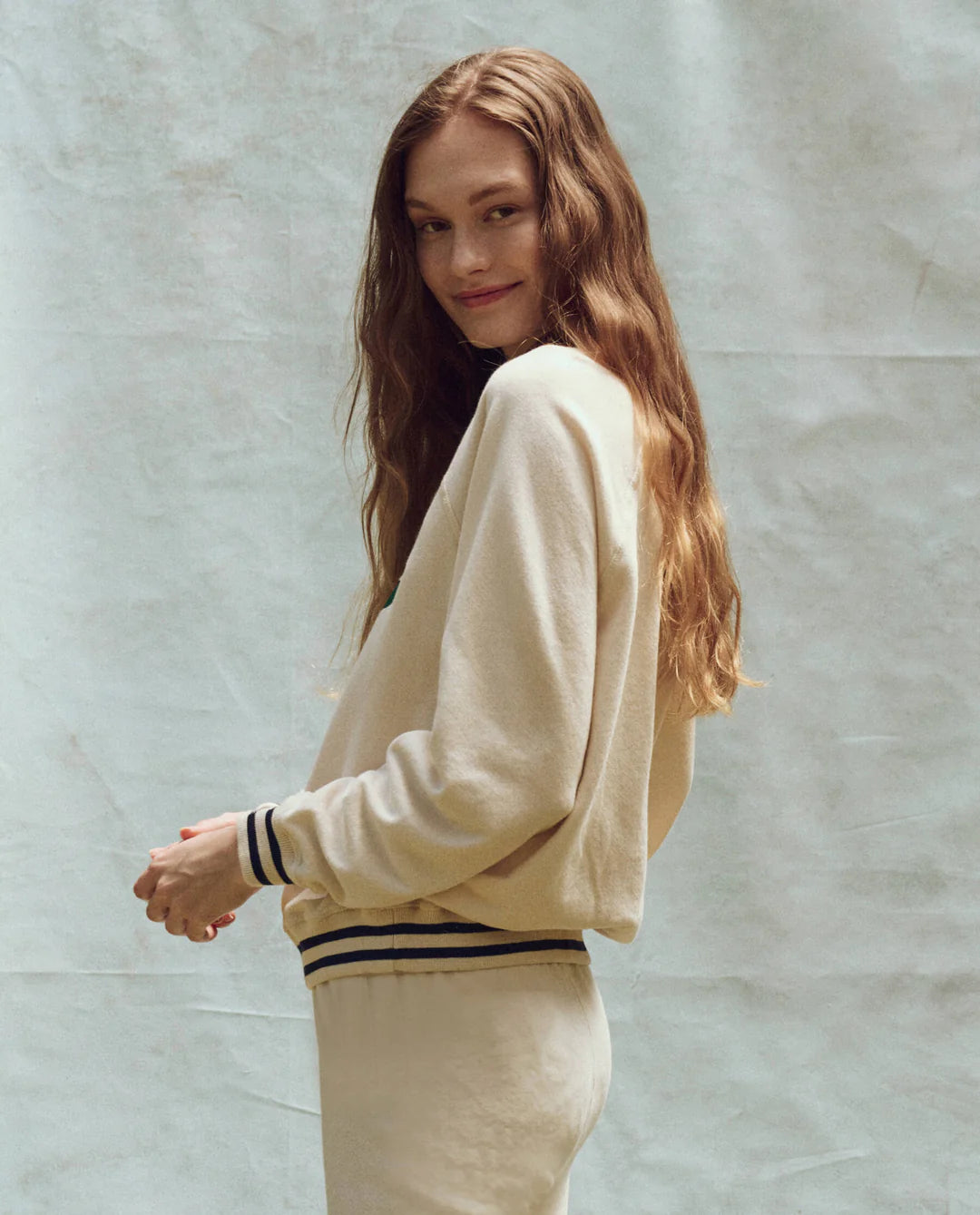 A person with long hair smiles while looking over their shoulder, dressed in The Great Inc.&#39;s oversized Varsity College Sweatshirt w/ Crest Graphic and matching pants. The soft cotton terry fabric and light colors beautifully complement the textured background.