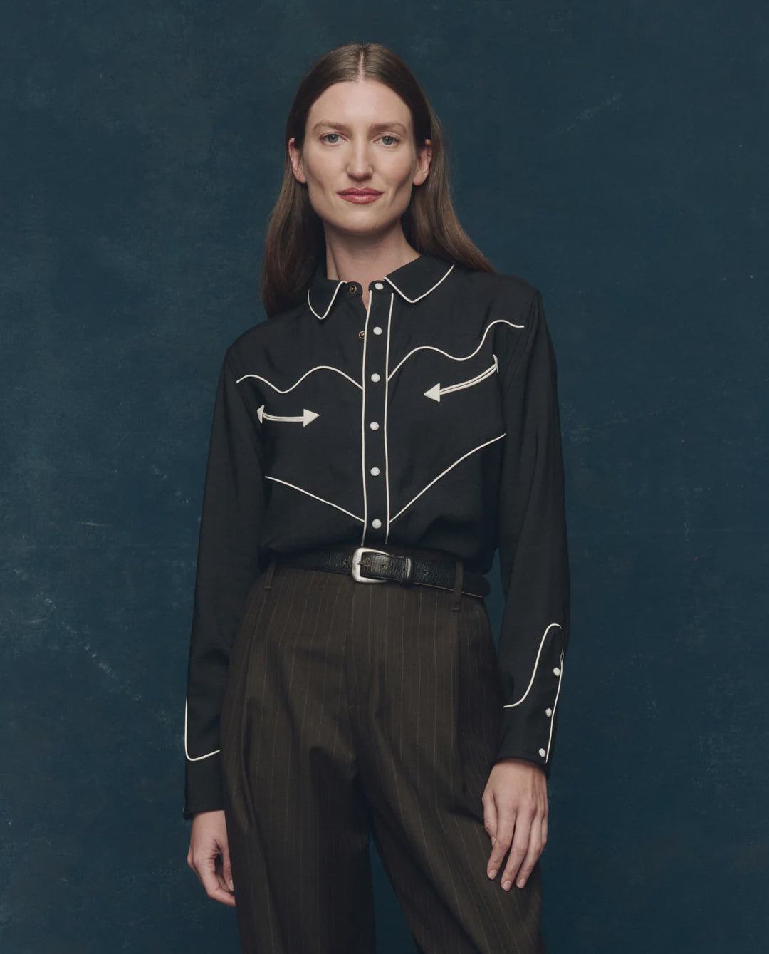 A woman with straight brown hair stands against a dark blue background, smiling softly at the camera while wearing The Western Shirt by The Great Inc.—a black shirt with white piping, arrow details, and pearl snaps—paired with high-waisted dark pants and a belt.