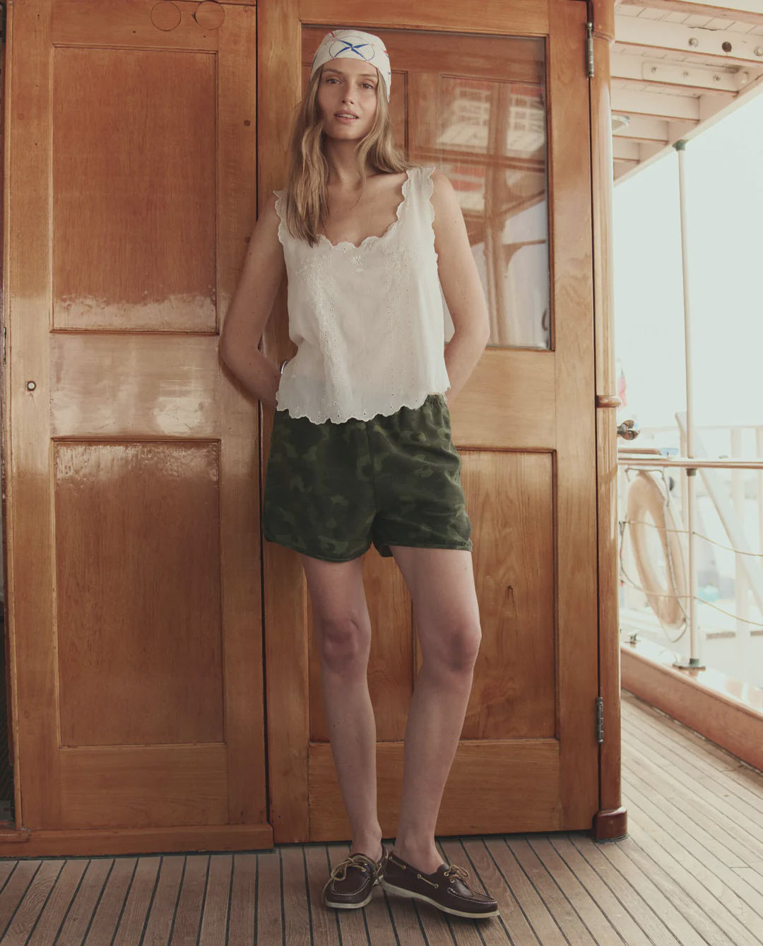 A woman on a boat deck in front of wooden doors wears The Dolphin Short by The Great Inc., a white scalloped tank top, brown boat shoes, and a white headscarf with blue trim.