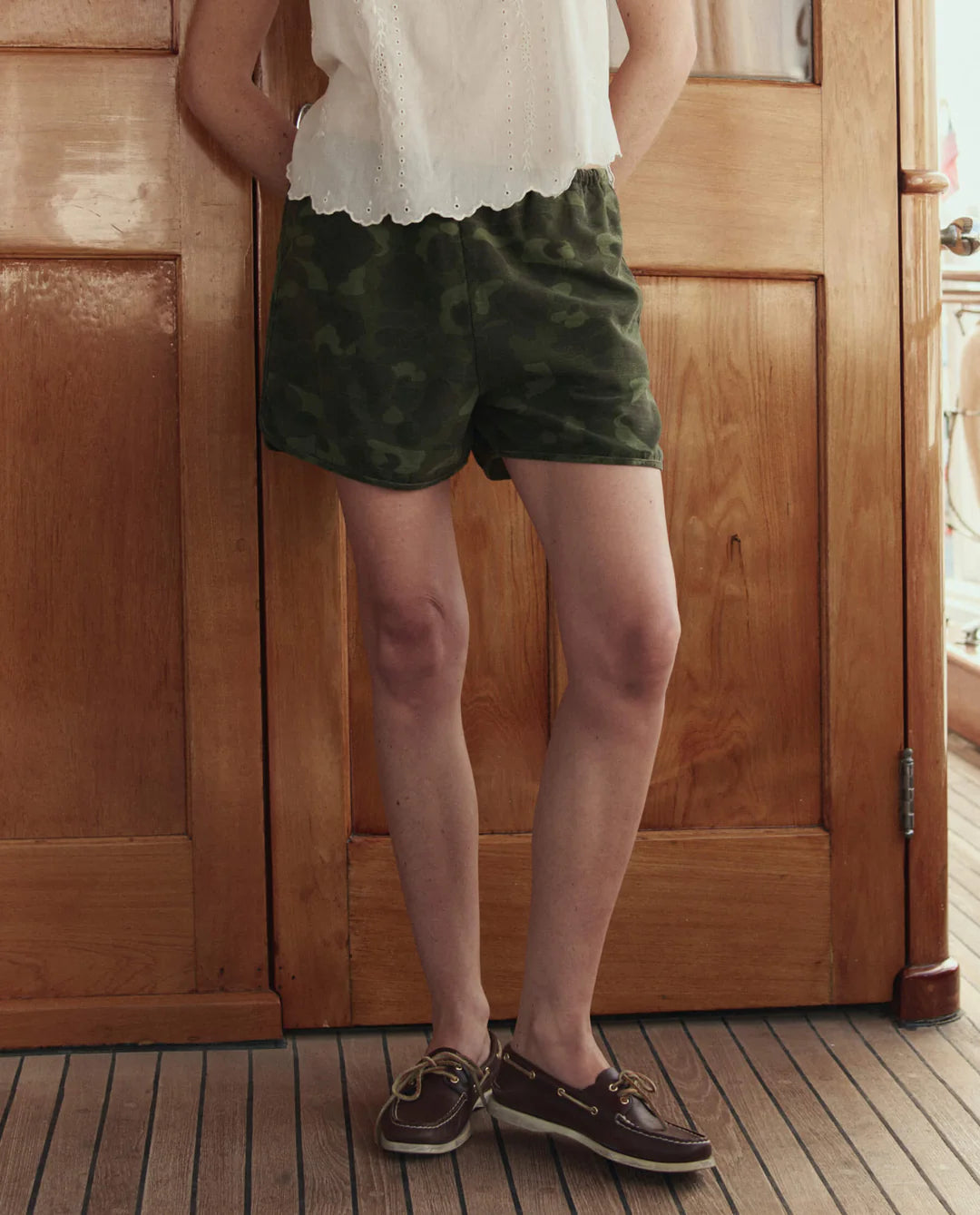A person stands indoors on a wooden floor, wearing a white sleeveless blouse, The Great Inc. Dolphin Short in green camo cotton sateen, and brown boat shoes. Their head is not visible in the image.