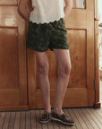 A person stands indoors on a wooden floor, wearing a white sleeveless blouse, The Great Inc. Dolphin Short in green camo cotton sateen, and brown boat shoes. Their head is not visible in the image.