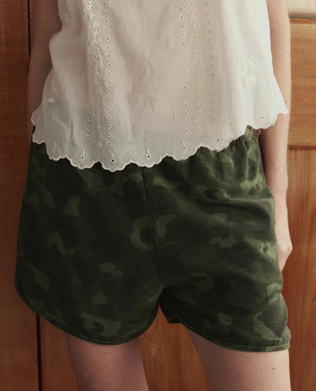 A person wearing a white, sleeveless, embroidered blouse and The Dolphin Short by The Great Inc. stands in front of a wooden door.