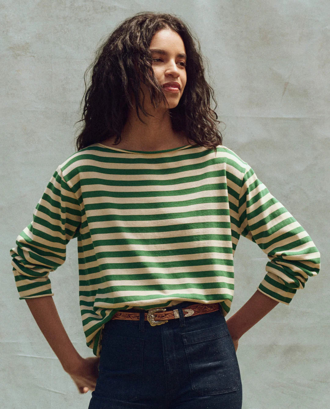 A person with long, wavy hair models The Great Inc.'s Sailor Sweater in green and white stripes, paired with dark jeans and a brown belt. Standing with hands on hips against a neutral backdrop, they capture a nautical uniform vibe while looking to the side.