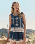 A woman stands outdoors in a desert landscape, wearing The Sparrow Tank by The Great Inc., a vintage-inspired blue sleeveless top with white embroidery and cotton crochet lace trim. Cacti are visible against the clear sky.