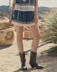 Outdoors on sandy ground, a person wears The Sparrow Short by The Great Inc.—vintage-inspired blue shorts with white lace trim and black cowboy boots—amid rocks, cacti, and desert plants. Their upper body is not visible.