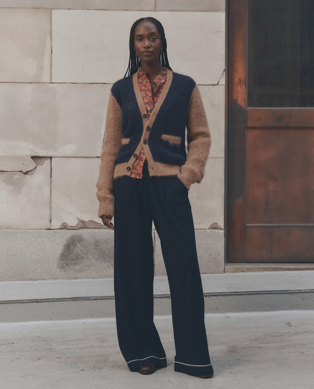 Patterned shirt and wide-leg pants by The Great Inc., modeled by a stone and metal wall, relaxed modern style.