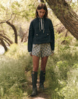 A person stands on a narrow forest path surrounded by greenery, wearing The Great Inc.'s Trailhead Pullover featuring a dark hood and front pocket, along with patterned shorts and knee-high boots. The scene is peaceful with trees and grass in the background.