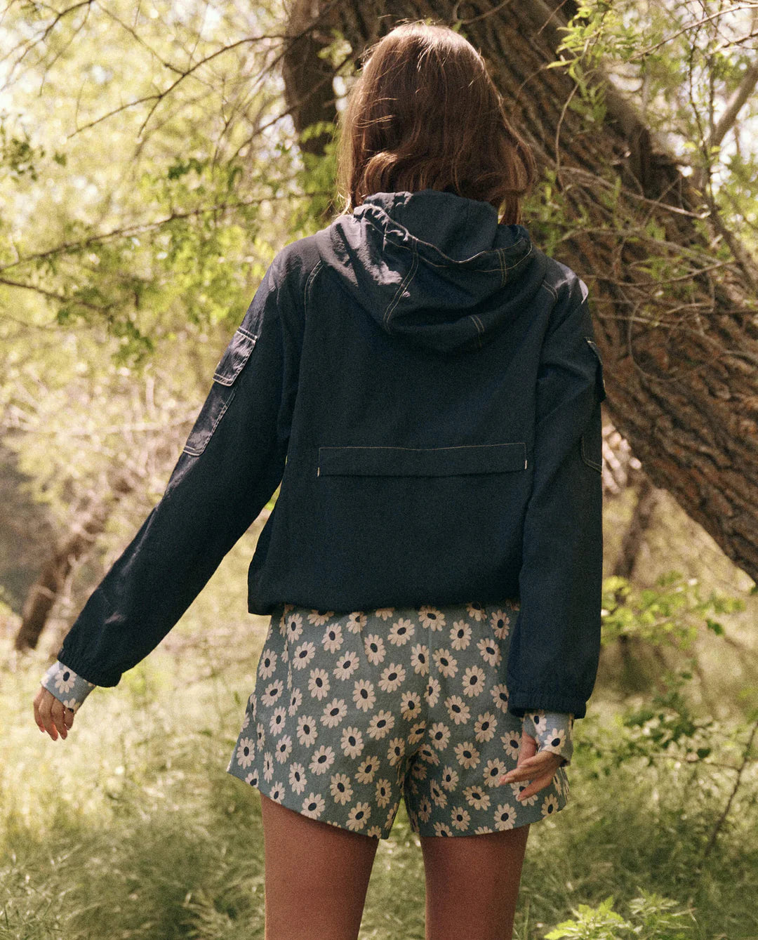 Clad in The Great Inc.'s dark, water-repellent Trailhead Pullover and floral shorts, a person stands in a sunlit, grassy area surrounded by trees. The scene captures their back as they face away, framed by branches and foliage.