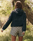 Clad in The Great Inc.'s dark, water-repellent Trailhead Pullover and floral shorts, a person stands in a sunlit, grassy area surrounded by trees. The scene captures their back as they face away, framed by branches and foliage.