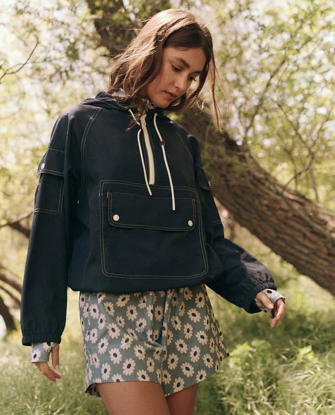 In a sunny garden, a woman wearing The Great Inc.'s water-repellent Trailhead Pullover and floral-patterned shorts stands amid lush greenery, creating a serene and contemplative atmosphere as she looks down.