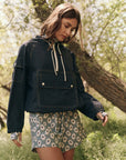 In a sunny garden, a woman wearing The Great Inc.'s water-repellent Trailhead Pullover and floral-patterned shorts stands amid lush greenery, creating a serene and contemplative atmosphere as she looks down.