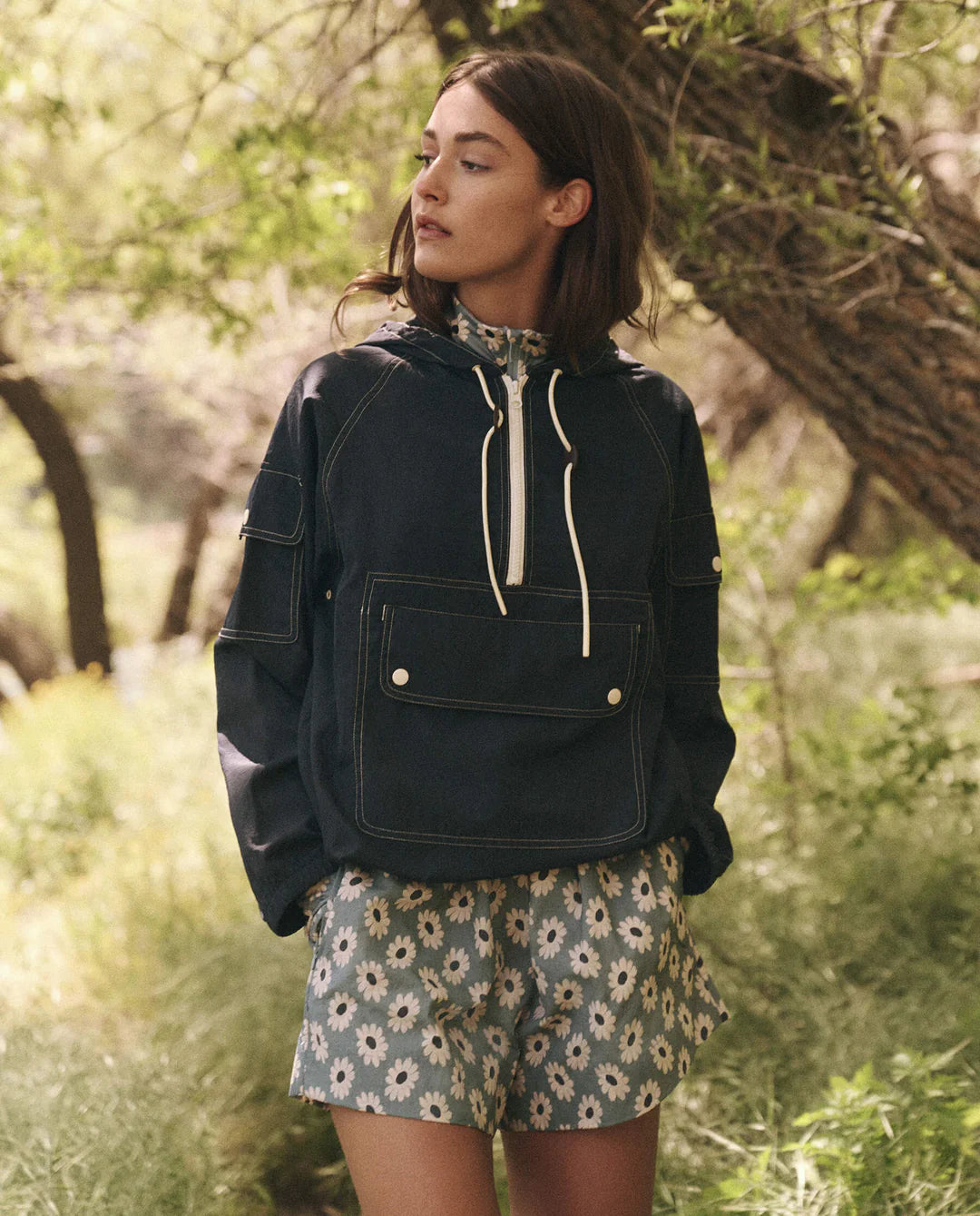 Amid trees and greenery, a woman in The Great Inc.'s Trailhead Pullover with a front pocket and floral shorts gazes sideways, harmonizing with the serene natural surroundings that enhance her technical apparel.