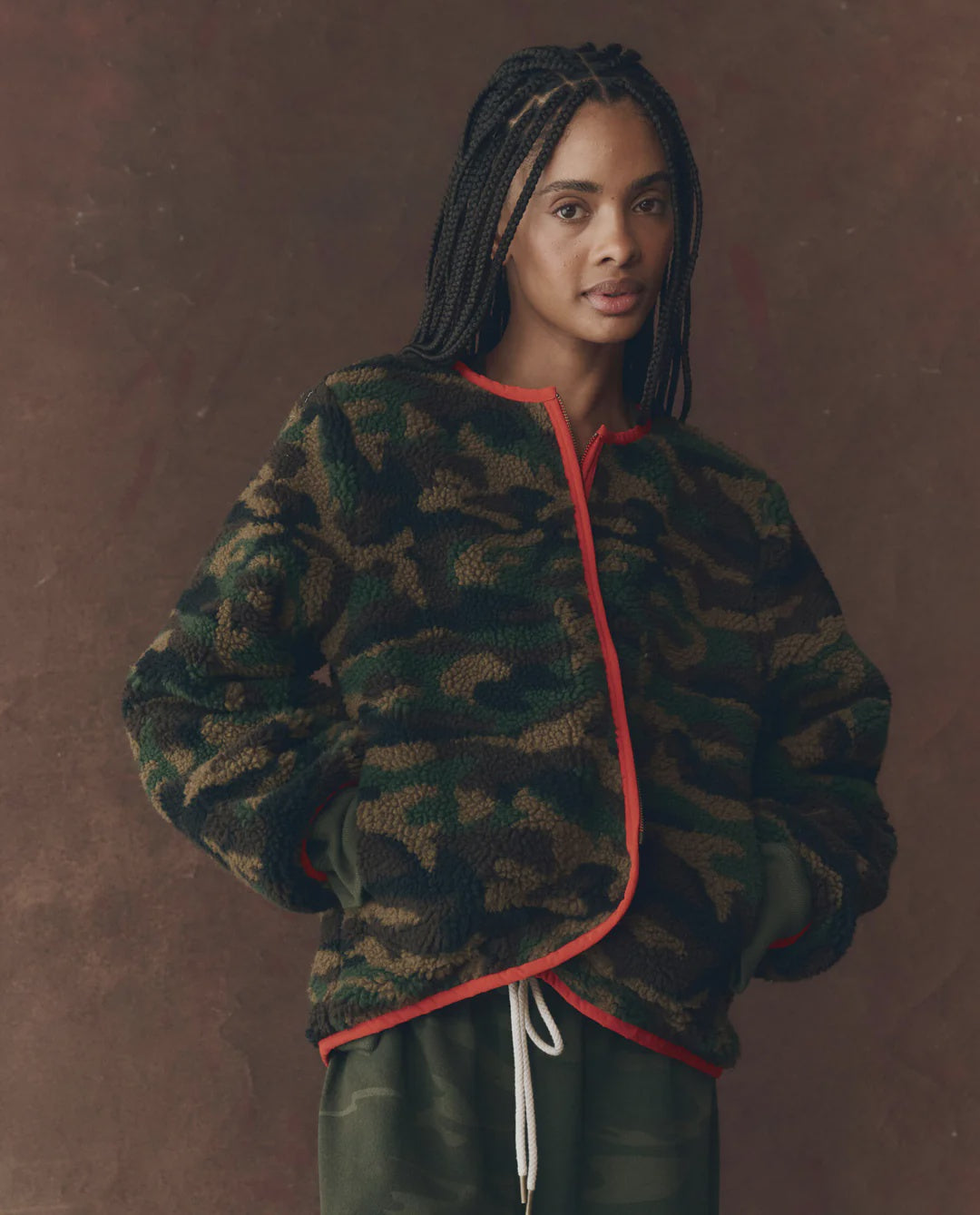 A woman with long braids wears The Great Inc. Reversible Plush Bomber, a vintage-style camo fleece jacket with red trim, over green pants against a brown textured background.