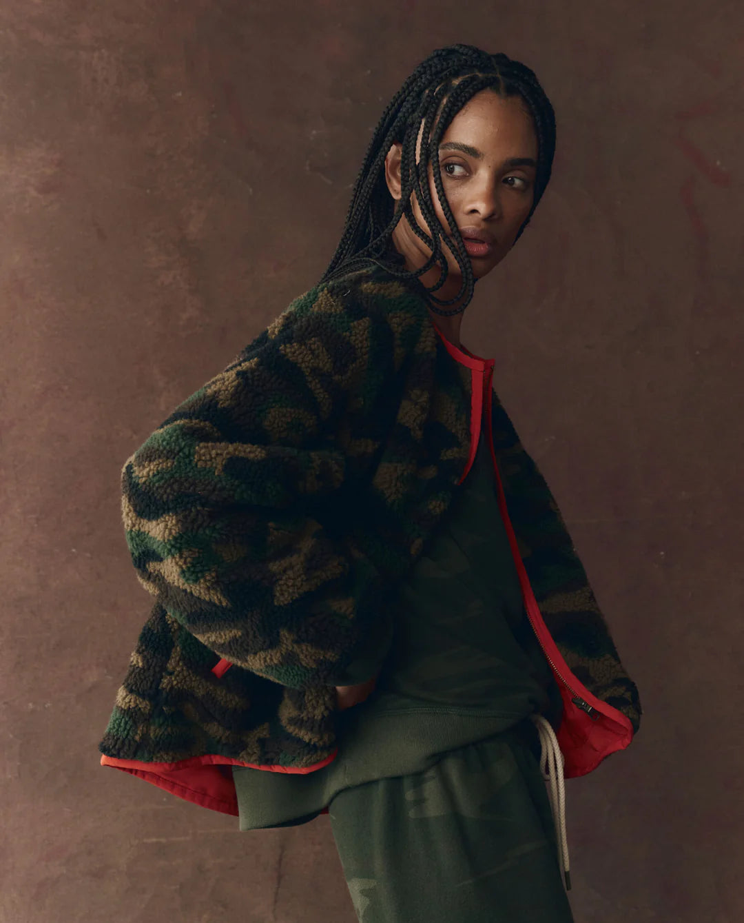 A person with braided hair poses before a brown backdrop, wearing The Great Inc. Reversible Plush Bomber with vintage camo fleece, red lining, and green clothing underneath.
