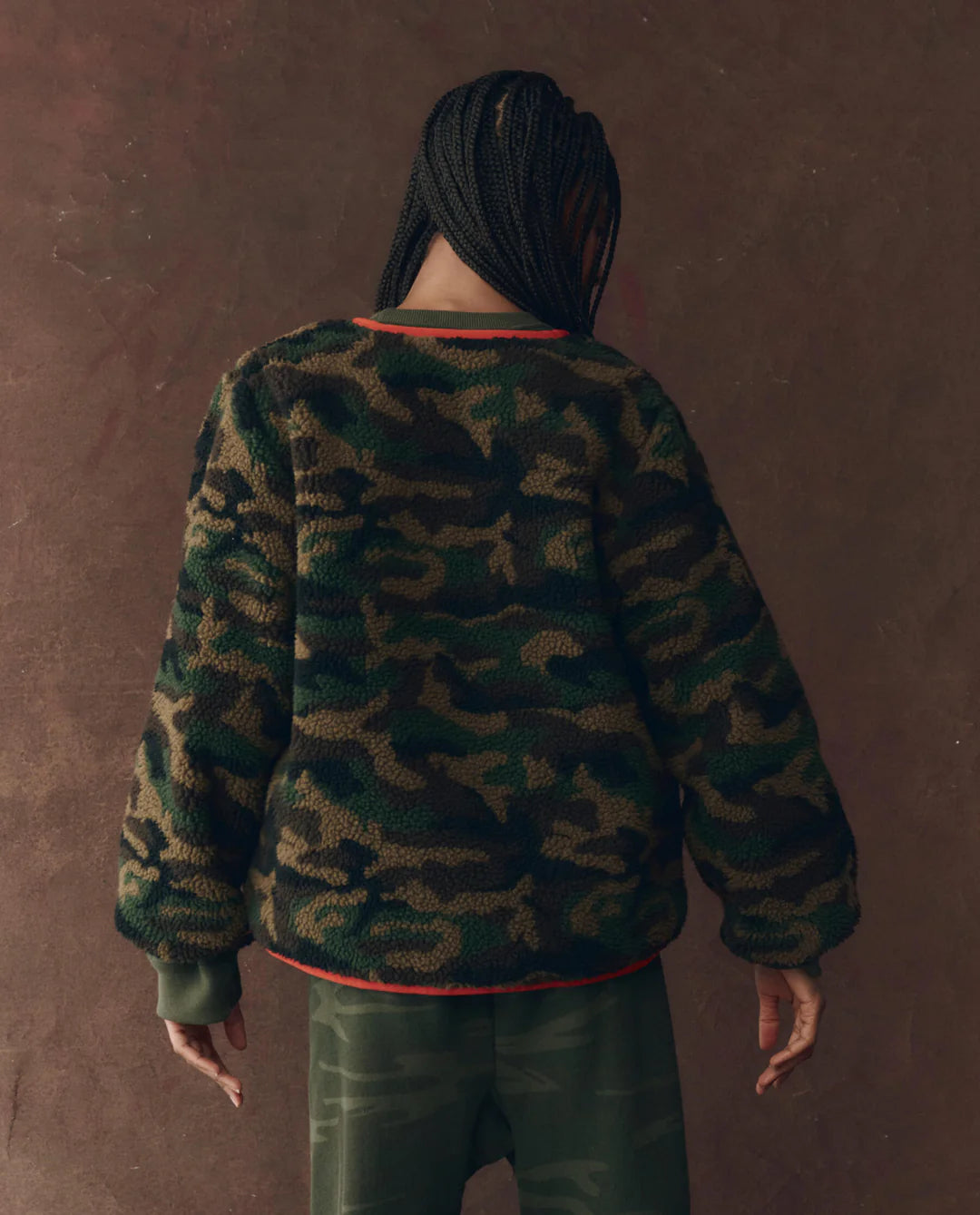A person with braided hair faces away, wearing The Great Inc. Reversible Plush Bomber in camo fleece against a textured brown background.