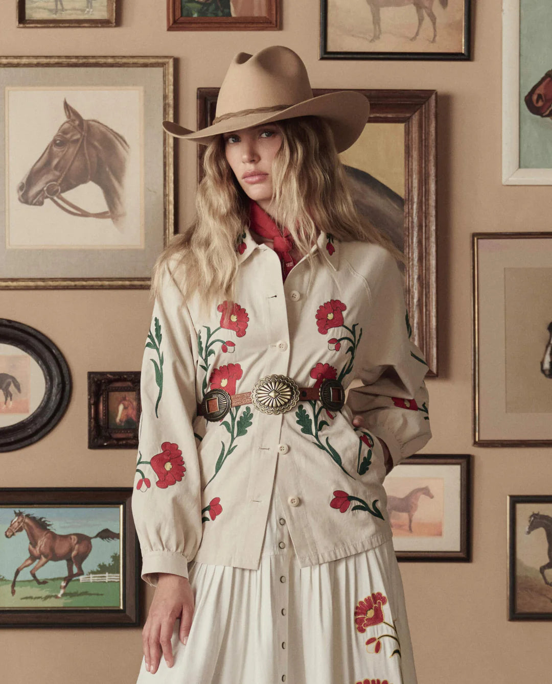 A woman wearing a beige embroidered outfit with red flowers and The Great Inc. Leather Concho Belt stands before a wall of framed horse paintings, sporting a tan cowboy hat and looking at the camera.