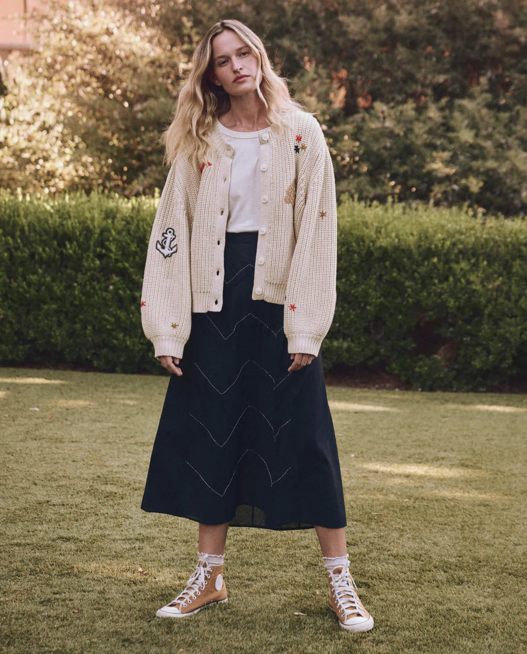 Amid greenery, a person stands on grass, wearing The Collegiate Cardigan by The Great Inc., paired with a white top and long black skirt with zigzag patterns. Tan high-top sneakers complete the ensemble.