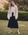 Amid greenery, a person stands on grass, wearing The Collegiate Cardigan by The Great Inc., paired with a white top and long black skirt with zigzag patterns. Tan high-top sneakers complete the ensemble.