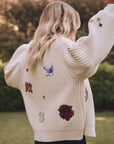 A person with long, blonde hair is wearing The Collegiate Cardigan by The Great Inc., a cream-colored knit adorned with colorful patches featuring hand-embroidered animals and collegiate graphics. They stand on a grassy lawn with trees in the background.