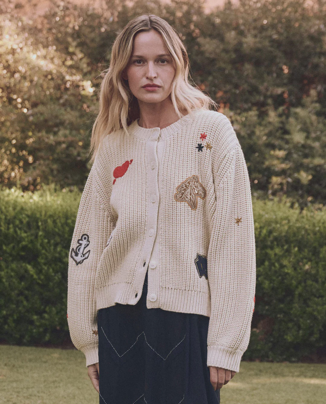 Wearing The Great Inc.'s Collegiate Cardigan, featuring hand embroidery and collegiate graphics with anchor and horse patches, a person stands outdoors surrounded by lush green foliage.