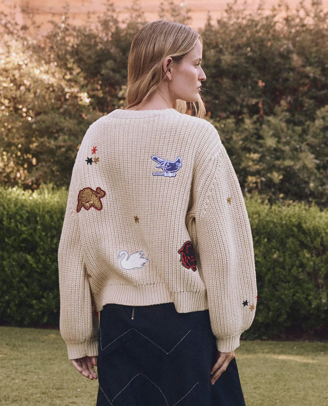 A woman with long blonde hair stands outdoors, wearing The Great Inc.'s cream-colored Collegiate Cardigan featuring collegiate graphics and animal patches like a blue bird and red bear. She pairs it with a dark skirt against a lush green backdrop.