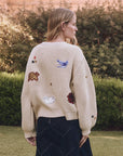 A woman with long blonde hair stands outdoors, wearing The Great Inc.'s cream-colored Collegiate Cardigan featuring collegiate graphics and animal patches like a blue bird and red bear. She pairs it with a dark skirt against a lush green backdrop.