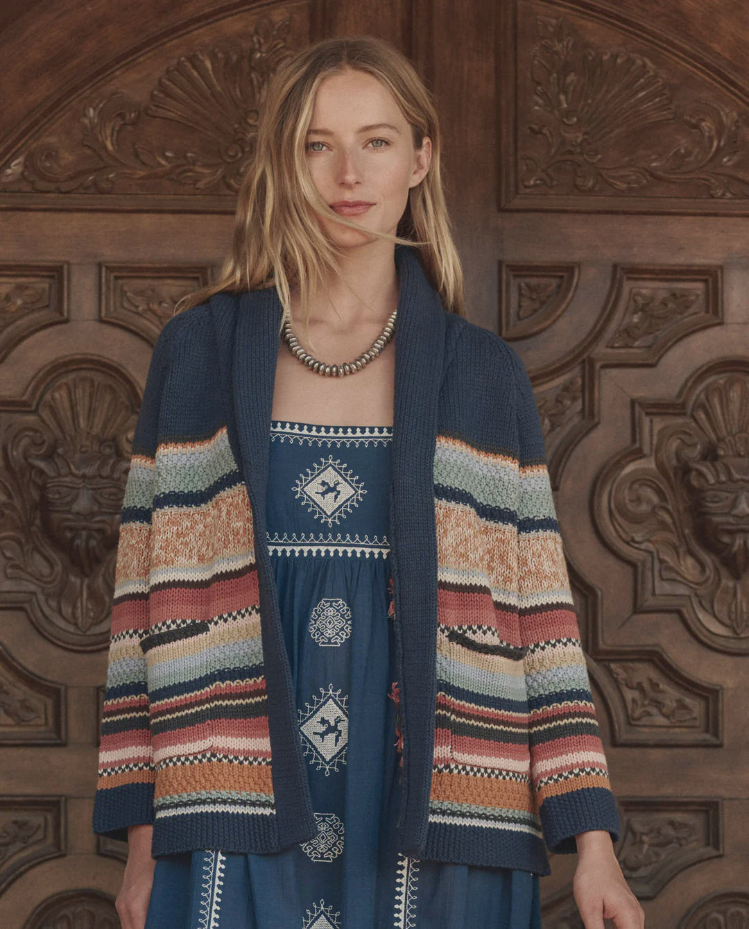 A woman with long blonde hair stands before an ornate wooden door in a blue patterned dress and The Great Inc.'s Mountain Lodge Cardigan, a chunky knit with navy, orange, cream, and red stripes. She accessorizes with a beaded necklace and serene expression.