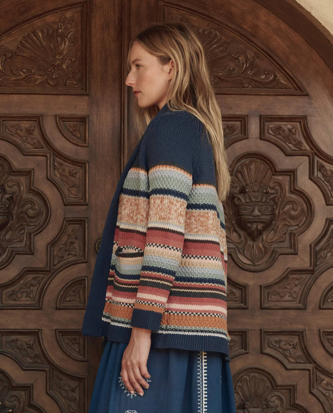 A woman in profile stands before ornate wooden doors, wearing The Mountain Lodge Cardigan by The Great Inc., a Southwestern-inspired piece with blue, orange, and cream horizontal stripes over a blue dress.
