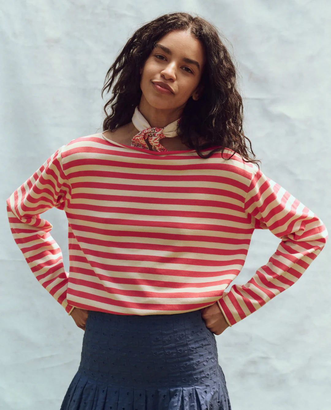 A person with long, dark hair is smiling and wearing The Great Inc.'s Sailor Sweater, a cotton yarn red and white striped long-sleeve shirt. They pair it with a dark blue skirt and a beige scarf with a red pattern. The background is plain and light.