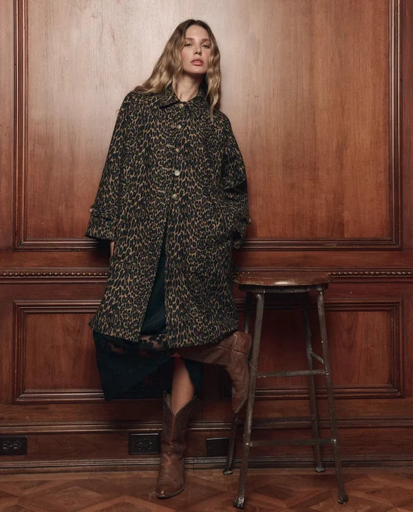 The Great Inc. trench coat in neutral tones styled with brown boots, set against a warm wood-paneled interior wall.