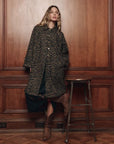 The Great Inc. trench coat in neutral tones styled with brown boots, set against a warm wood-paneled interior wall.