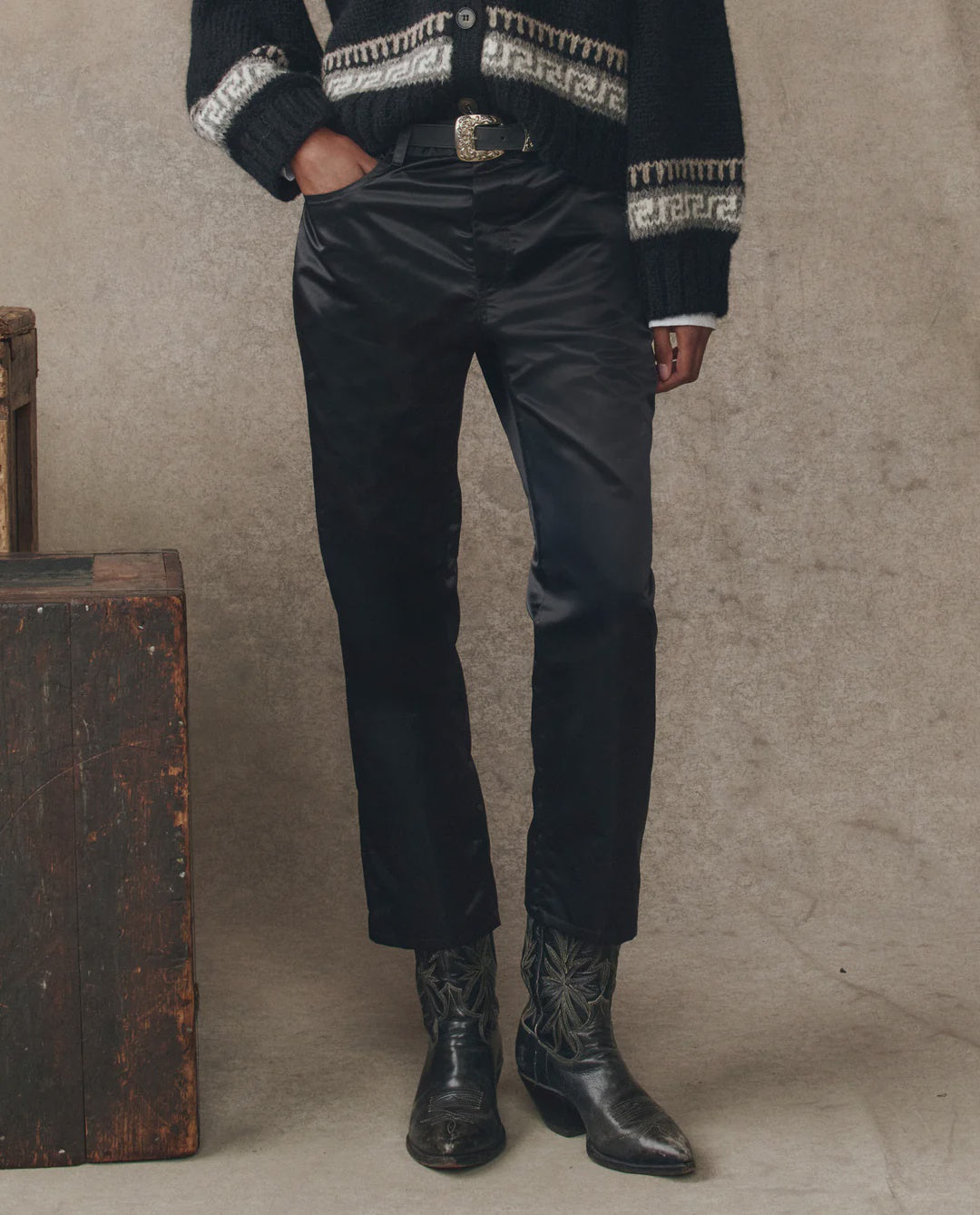 A person stands indoors with hands in their pockets, wearing The Satin Wayne cropped pants by The Great Inc., a black sweater with a white geometric pattern, a decorative-buckle belt, and black stitched cowboy boots.