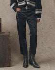 A person stands indoors with hands in their pockets, wearing The Satin Wayne cropped pants by The Great Inc., a black sweater with a white geometric pattern, a decorative-buckle belt, and black stitched cowboy boots.