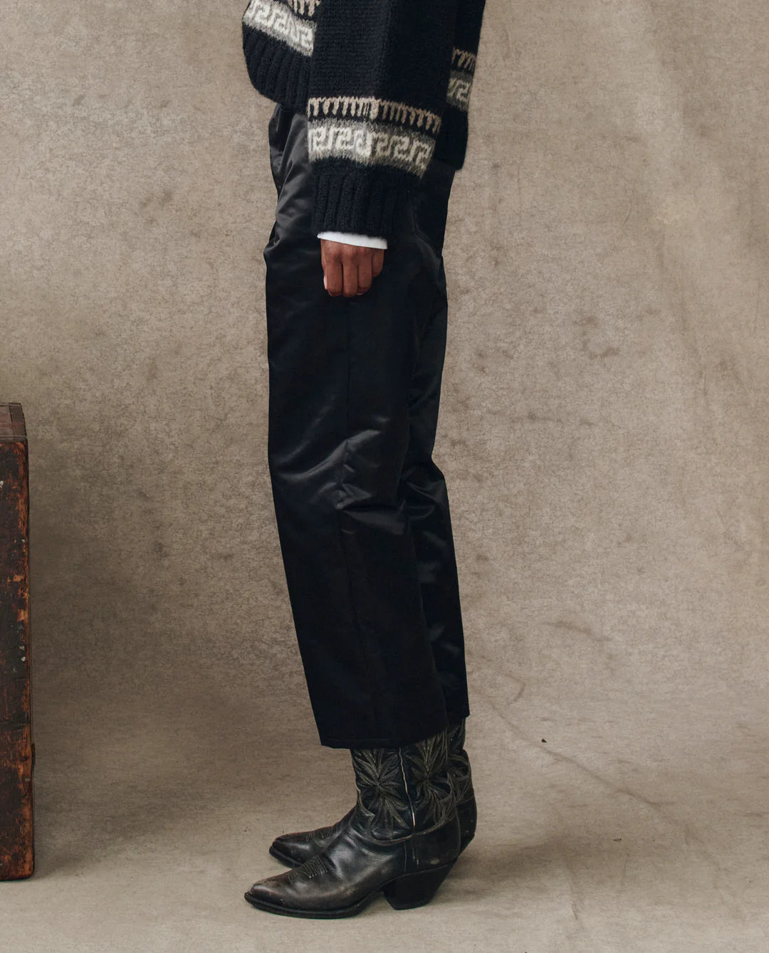 A person stands sideways wearing The Satin Wayne black slouchy satin pants by The Great Inc., paired with patterned black cowboy boots and a black sweater with white cuffs and designs, set against a neutral textured background.