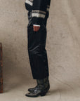 A person stands sideways wearing The Satin Wayne black slouchy satin pants by The Great Inc., paired with patterned black cowboy boots and a black sweater with white cuffs and designs, set against a neutral textured background.