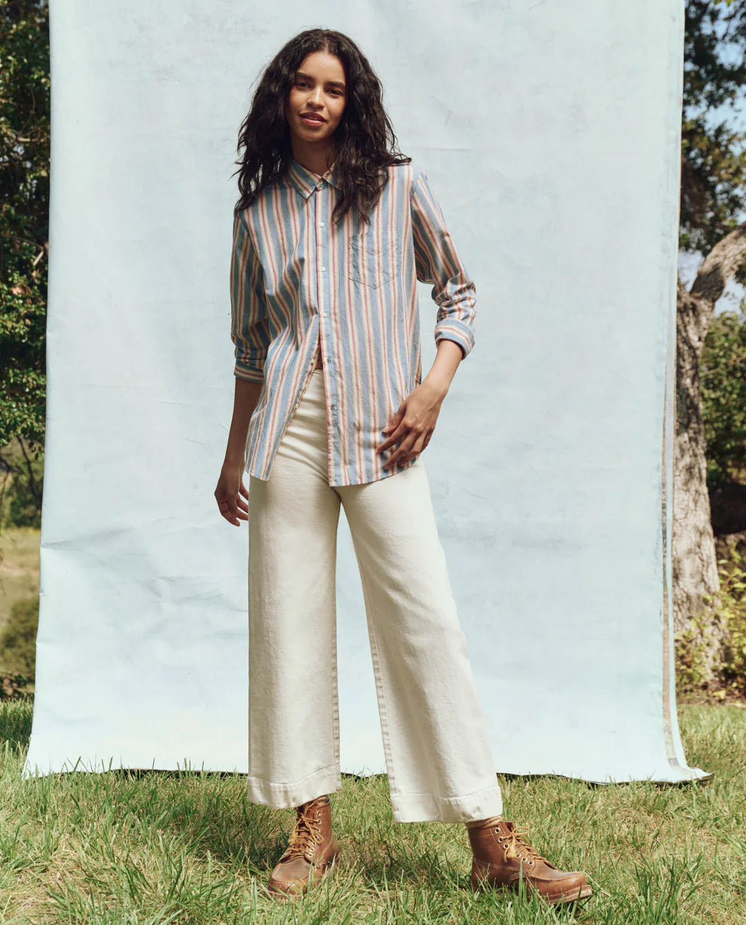 A person stands gracefully on grass with a light blue backdrop, wearing The Great Inc.'s Post Top, featuring menswear shirting stripes and a button-up collar. They're paired with white wide-leg pants and brown boots, surrounded by trees in a bright natural setting.