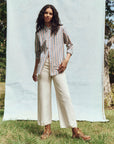 A person stands gracefully on grass with a light blue backdrop, wearing The Great Inc.'s Post Top, featuring menswear shirting stripes and a button-up collar. They're paired with white wide-leg pants and brown boots, surrounded by trees in a bright natural setting.