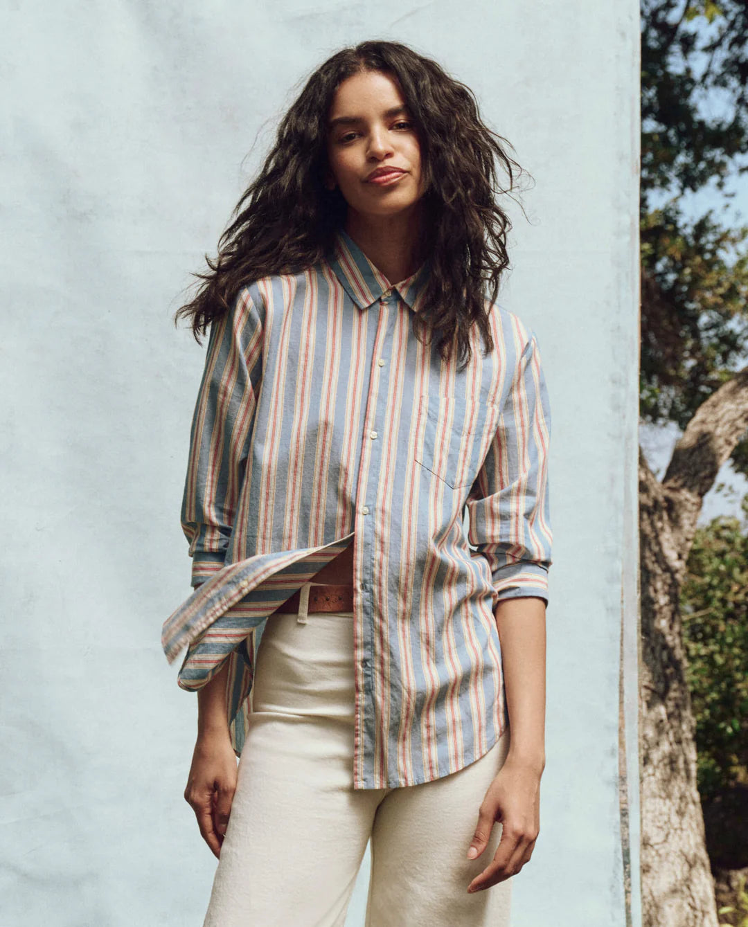 A person with long, wavy hair stands outdoors against a light blue background, wearing The Great Inc.'s The Post Top—a striped button-up shirt with a neat collar—and white pants. They smile slightly with one hand in their pocket, and a tree is visible in the background.