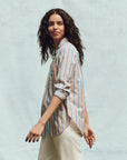 With long, wavy hair, a person wears The Great Inc.'s The Post Top in a relaxed fit and light-colored pants. They walk left with a slight smile against a plain, light background.