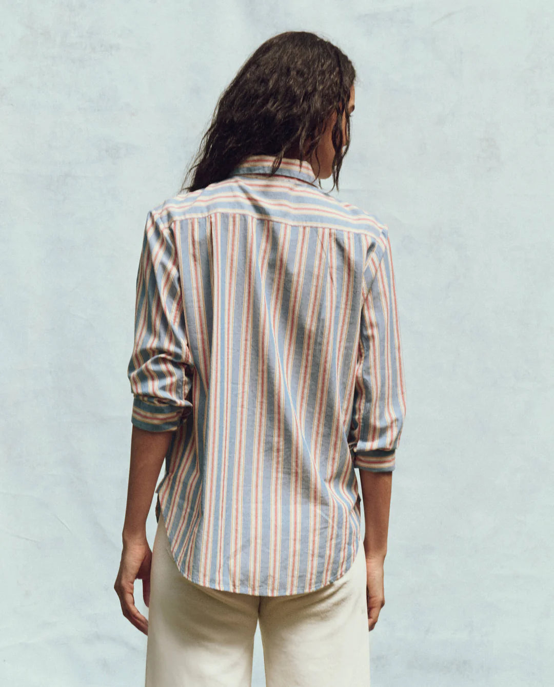 A person with long dark hair, standing with their back to the camera, is wearing The Post Top by The Great Inc. This relaxed-fit button-up features blue, orange, and white menswear shirting stripes. The sleeves are rolled up and it's paired with white pants against a light blue background.