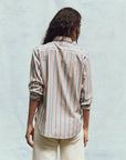 A person with long dark hair, standing with their back to the camera, is wearing The Post Top by The Great Inc. This relaxed-fit button-up features blue, orange, and white menswear shirting stripes. The sleeves are rolled up and it's paired with white pants against a light blue background.