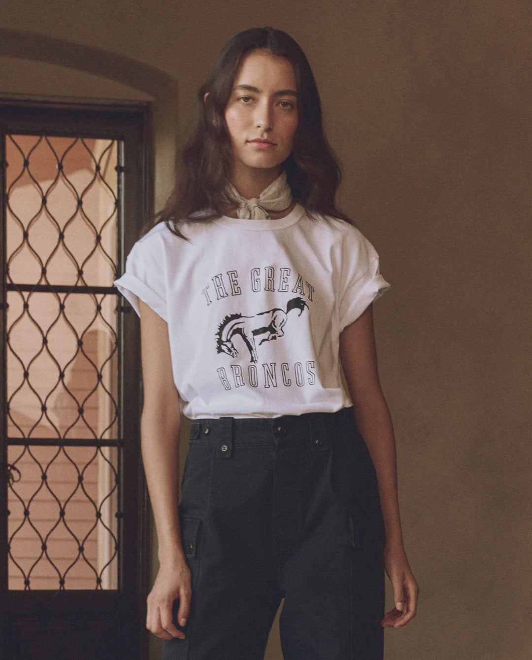 A person with long dark hair stands indoors near a window with a decorative metal grille, wearing The Great Inc.’s vintage-inspired "The Boxy Crew w/ Bucking Bronco" 100% cotton white t-shirt, black pants, and a light scarf around their neck.