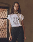 A person with long dark hair stands indoors near a window with a decorative metal grille, wearing The Great Inc.’s vintage-inspired "The Boxy Crew w/ Bucking Bronco" 100% cotton white t-shirt, black pants, and a light scarf around their neck.